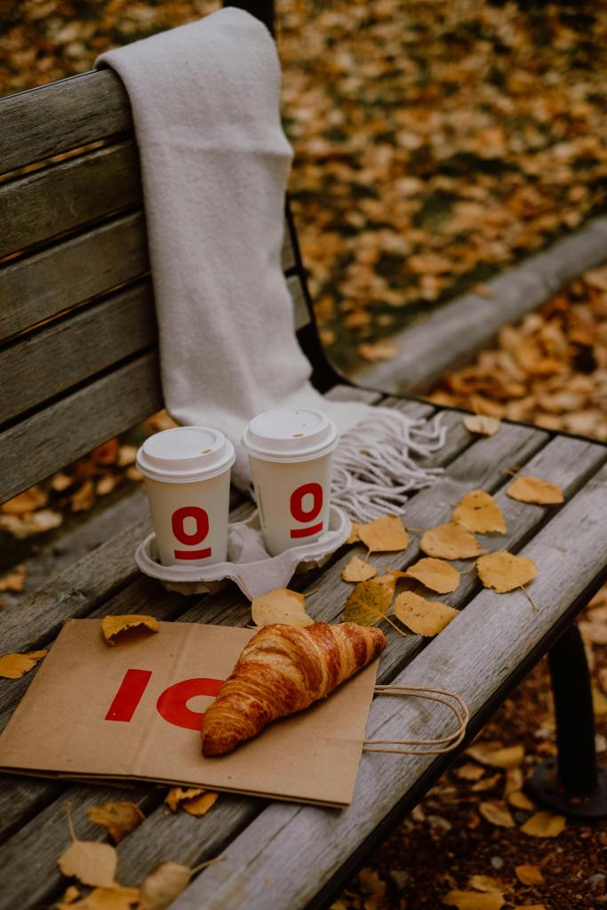 A cozy park bench covered with golden autumn leaves features a warm white scarf draped over the back, two cups of coffee, and a croissant on a paper bag, creating a comforting fall scene.