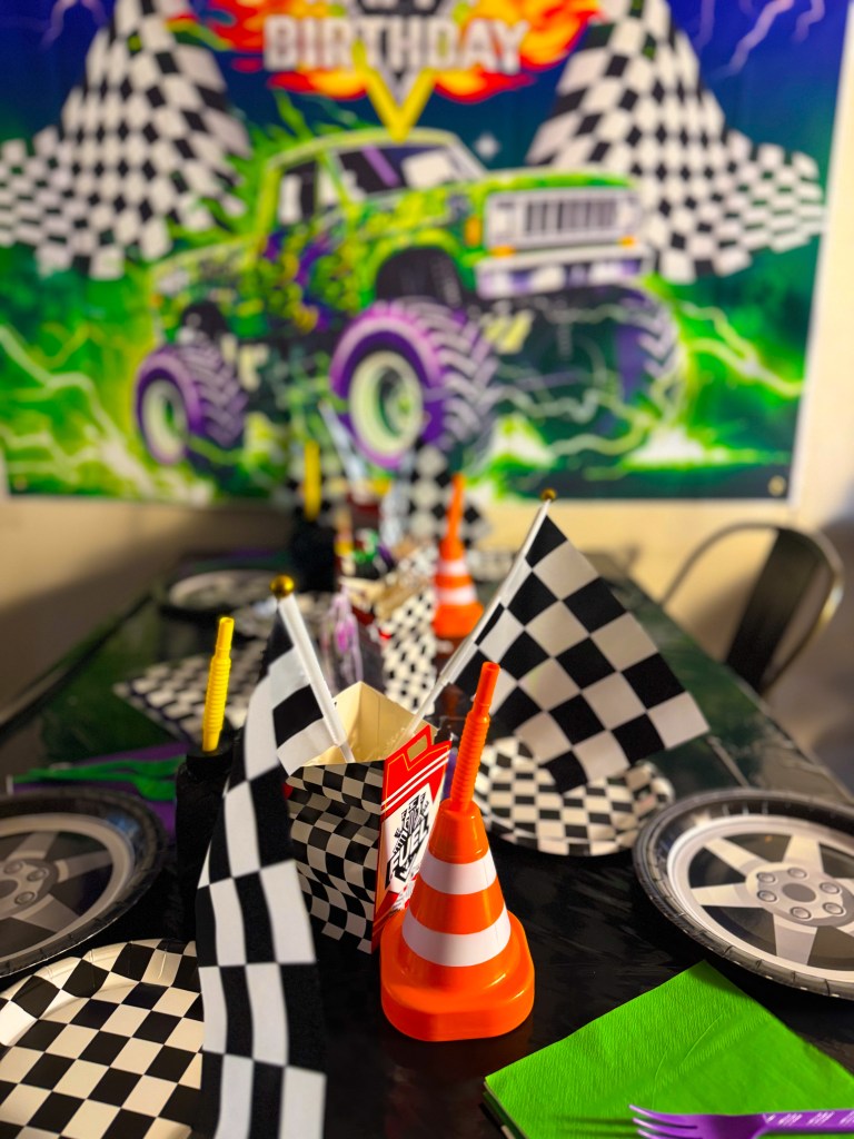Monster truck birthday party table decorated with checkered racing flags, tire themed plates, traffic cone cups, and Monster Jam truck banner in the background.