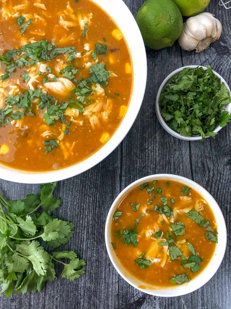 A warm bowl of soup featuring shredded chicken, corn, and a vibrant orange broth, garnished with fresh cilantro, alongside a small bowl of the same soup and various herbs and limes.