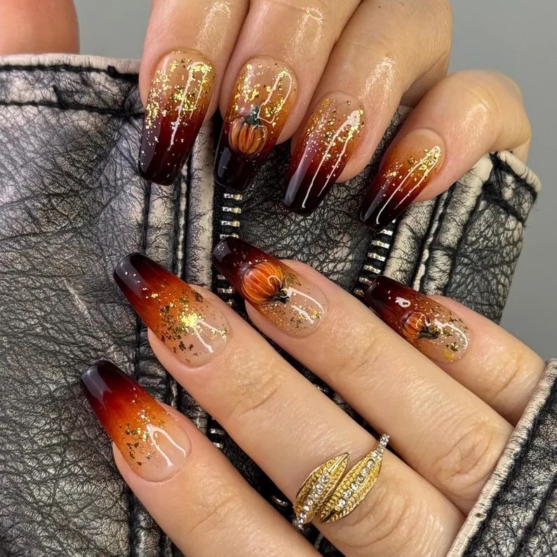 Elegant long nails featuring an ombré design with deep burgundy tips, golden glitter accents, and delicate pumpkin art, set against a textured leather background.