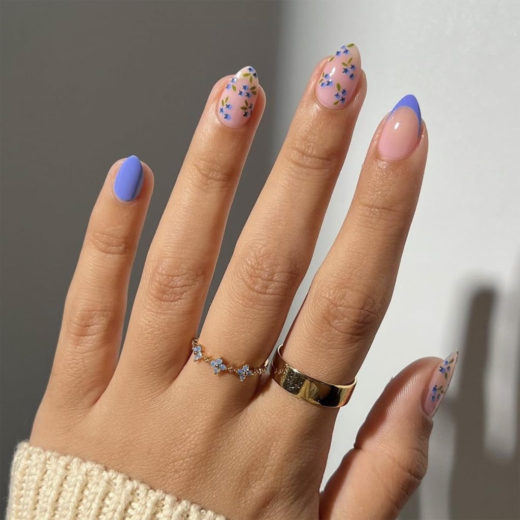 A hand displaying stylish nails with floral designs and vibrant colors, featuring a mix of pastel blue and beige. Two rings are worn on the fingers, adding elegance to the look.