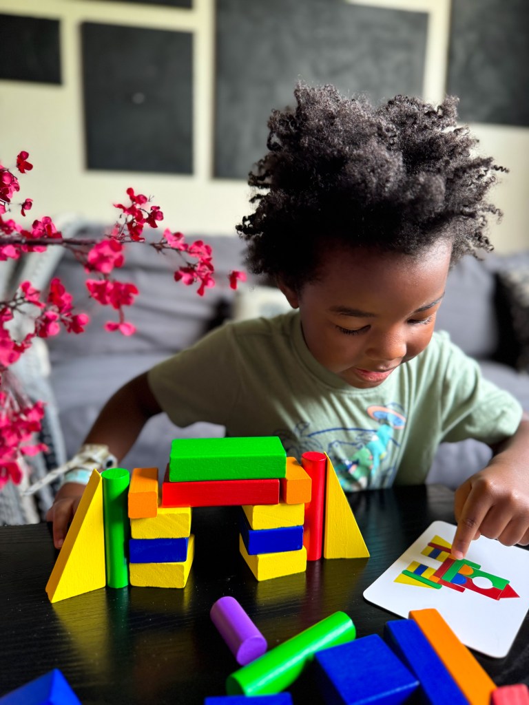 A young child with curly hair playing with colorful building blocks, focusing on a card with a design. The scene features a black couch in the background and a decorative plant nearby.