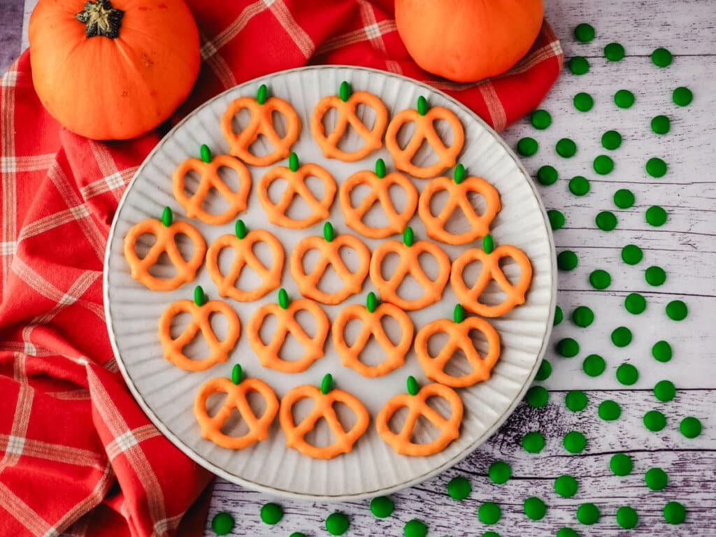 A plate of pumpkin-shaped pretzel treats decorated with orange and green candy, surrounded by small green candies and a red plaid cloth and two pumpkins.