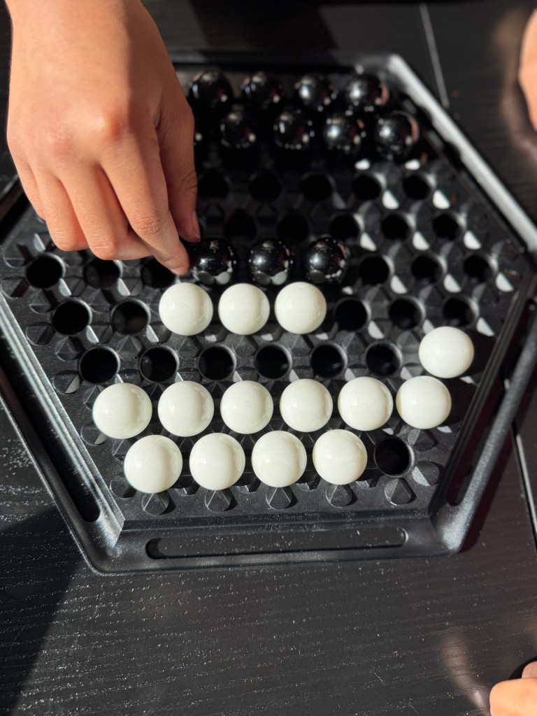 A close-up of hands playing the game Abalone on a hexagonal board, featuring black and white marbles arranged strategically.