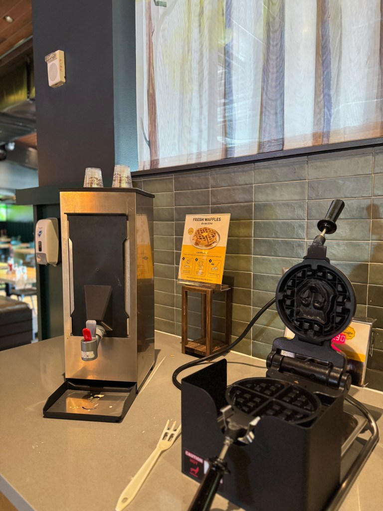 A waffle maker and syrup dispenser located in a breakfast area, with a sign promoting fresh waffles in the background.