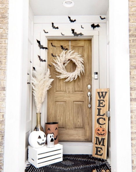 A cozy fall porch with a wooden door adorned with a natural wreath, accompanied by a 'WELCOME' sign and Halloween-themed decor, including a pumpkin and a ghost figurine. Black bats are attached to the wall, adding a playful touch to the festive atmosphere.