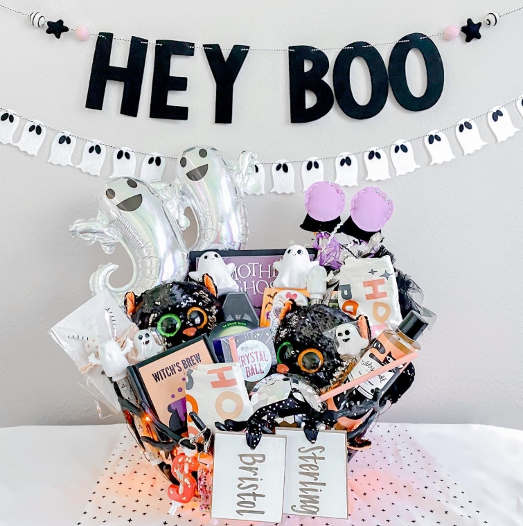 A decorative Halloween basket filled with themed treats, plushies, and accessories, featuring a 'Hey Boo' banner and ghost decorations.