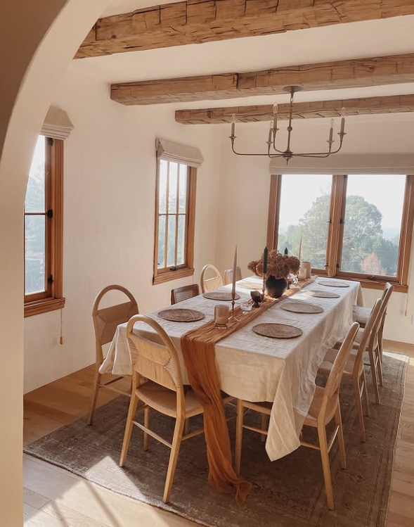 A warmly lit dining room featuring a long table set for a Thanksgiving gathering, adorned with beige linens, wooden placemats, candle holders, and a natural center arrangement, complemented by large windows with a scenic view.