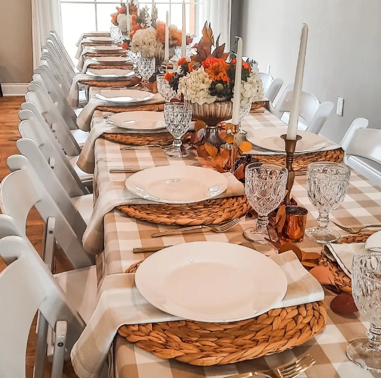 A beautifully styled Thanksgiving tablescape featuring a long table set with neutral-colored dishes, woven placemats, and crystal glassware. The table is adorned with fall-themed floral arrangements and candles, creating a warm and inviting atmosphere.