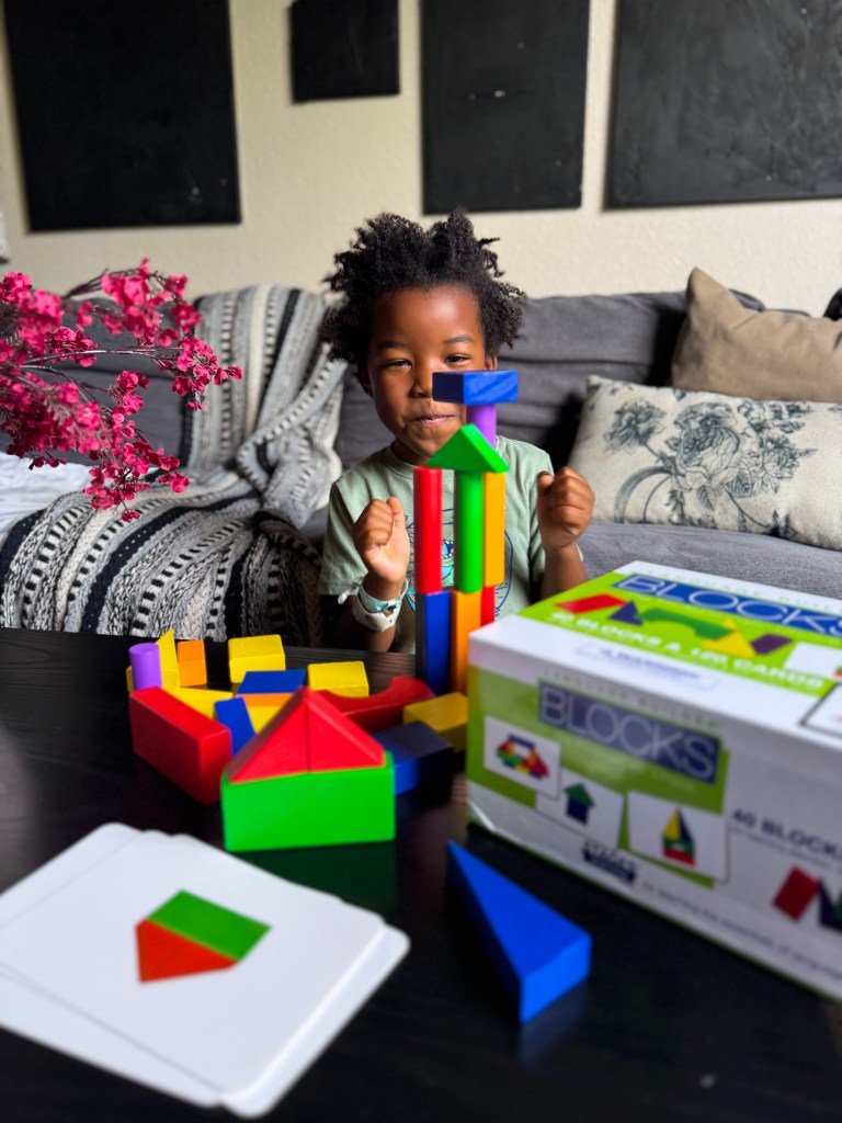A young child smiles while playing with colorful building blocks on a table, with a box of blocks beside them and various shapes scattered around.