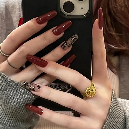 A close-up of hands with long, glossy burgundy nails featuring intricate nail art on some fingers, holding a phone, with a cozy sweater in the background.