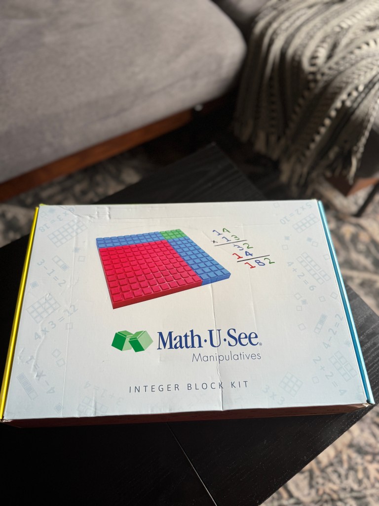 A close-up of the Math-U-See Integer Block Kit box, featuring colorful manipulatives and math equations, placed on a dark table with a sofa in the background.