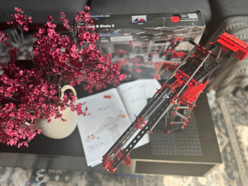 A top-down view of a completed Fischer Technik STEM kit model alongside its instruction manual, set on a black table with a vase of pink flowers in the background.