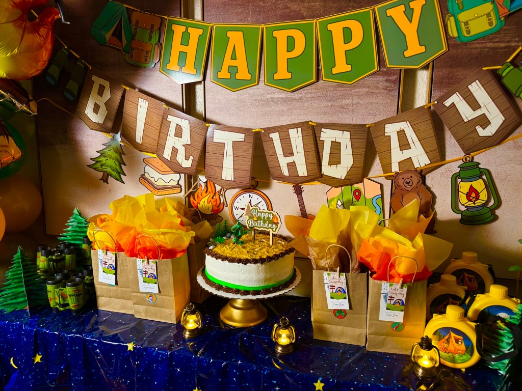 Kids camping party decorations with green binoculars, yellow canteens, and glowing lanterns on a gingham tablecloth.