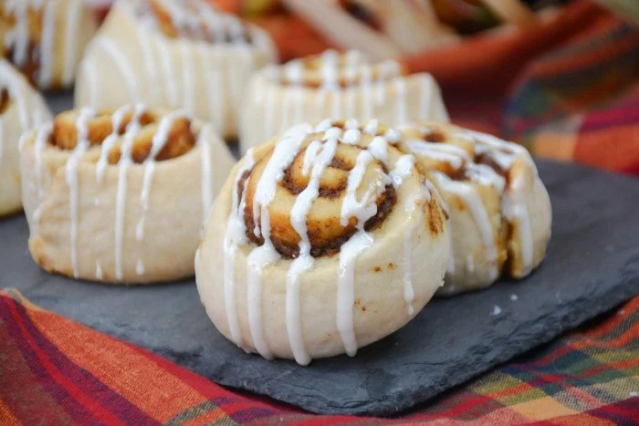Freshly baked pumpkin cinnamon rolls drizzled with cream cheese icing, presented on a slate platter with a colorful autumn-themed background.