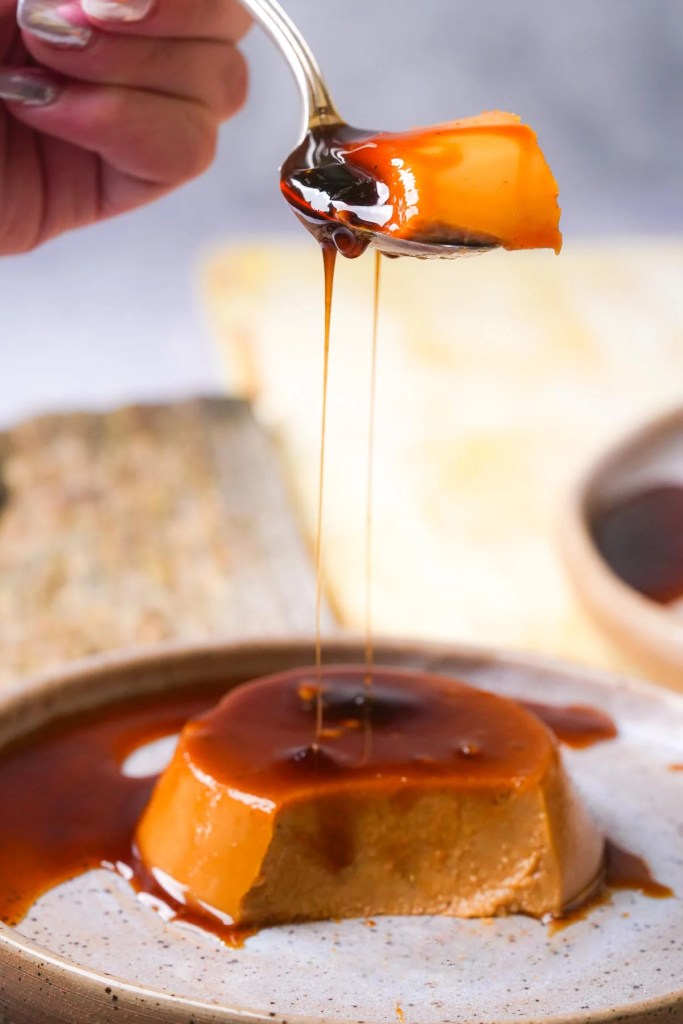 A spoon lifting a piece of pumpkin flan drizzled with caramel sauce, sitting on a plate.