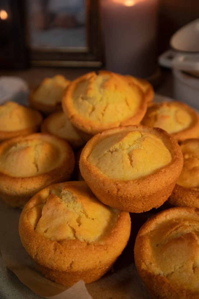 A pile of freshly baked vegan cornbread muffins on a wooden surface, with a soft glow from nearby candles in the background.