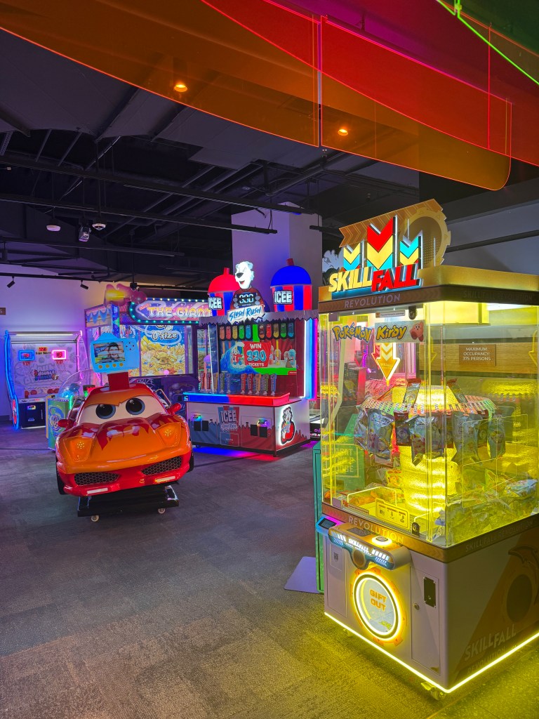 A vibrant and colorful arcade area featuring various games and attractions, including a car ride, ticket redemption machines, and flashing neon lights.
