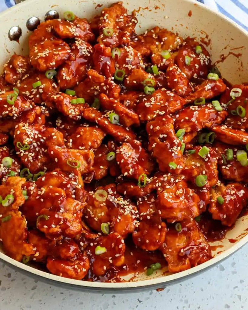 A close-up view of crispy fried chicken pieces coated in a glossy, spicy sauce garnished with sesame seeds and sliced green onions in a skillet.