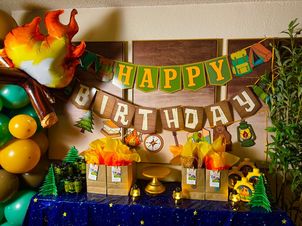 Indoor camping party dessert and favor table with birthday banner, goodie bags, and lantern decorations.