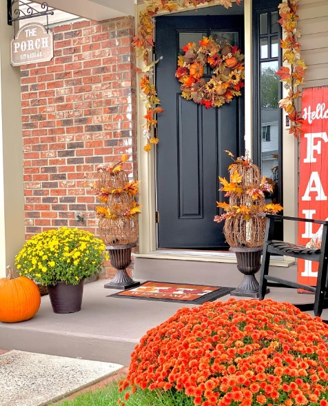 A cozy porch setup featuring a black front door adorned with a vibrant fall wreath. The entrance is decorated with colorful potted mums, seasonal foliage, and a rustic welcome sign, creating an inviting autumn atmosphere.
