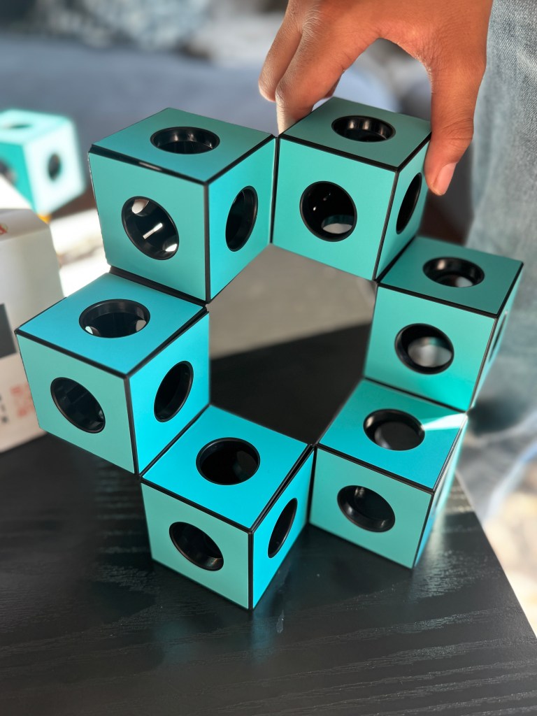 A person's hand manipulating a kinetic building set featuring turquoise cubes with circular openings, arranged in a circular formation on a dark surface.