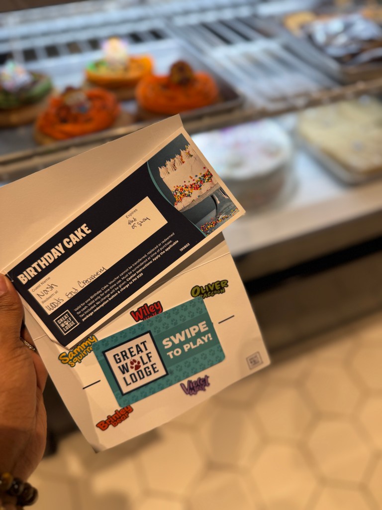 A hand holding a birthday cake pickup slip from Great Wolf Lodge in Grapevine, TX , with a colorful cake image on it, in front of a display case filled with various desserts.