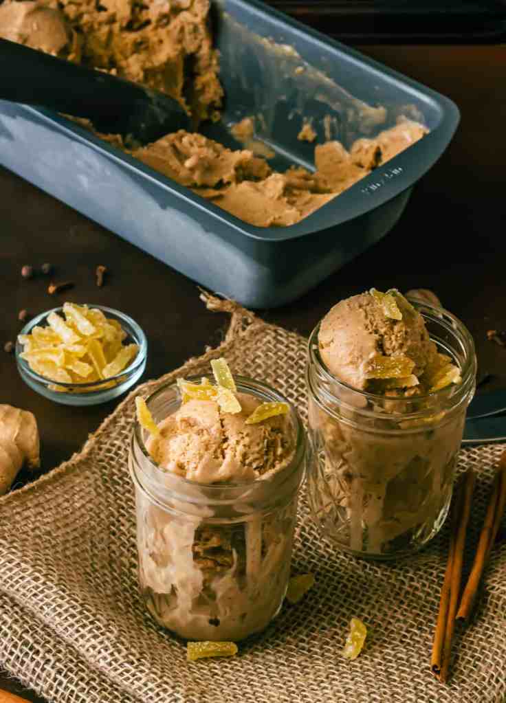 Two jars filled with creamy ginger pumpkin custard ice cream, topped with chopped candied ginger, placed on a rustic burlap surface alongside a scoop and cinnamon sticks.