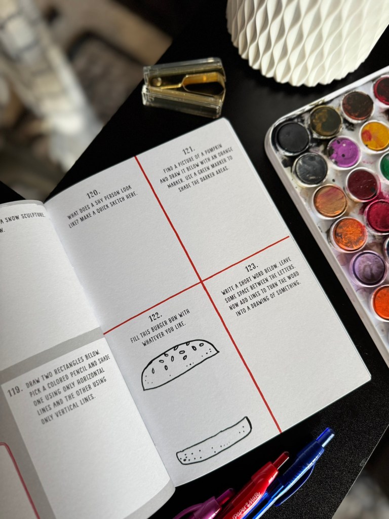 An open art book displaying several pages featuring drawing prompts, including instructions to sketch a snow sculpture and draw a burger bun, with colorful art supplies in the background.
