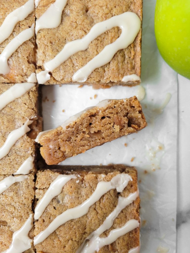 A plate of spiced apple blondies drizzled with white icing, with one piece partially cut out and a green apple beside it.