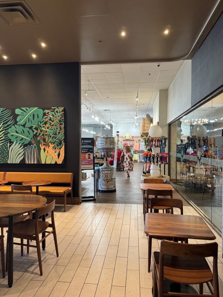 View of a cozy café space at Starbucks at Great Wolf Lodge in Grapevine, TX - with wooden tables and chairs, leading to a brightly lit retail area showcasing various merchandise such as swimwear and souvenirs, with a large Great Wolf Lodge display in the foreground.