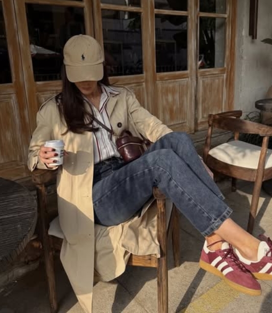 A woman sitting at a café wearing a beige trench coat, striped shirt, and dark denim jeans, holding a coffee cup. She's wearing a baseball cap and red sneakers.