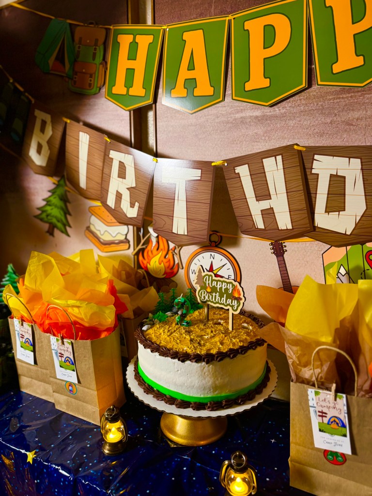 Camping birthday cake table with goodie bags, lanterns, and outdoor adventure party decorations.