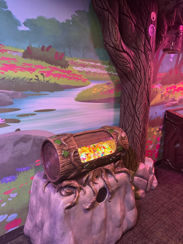 A colorful log-shaped treasure chest filled with bright, sparkling gems sits against a whimsical nature-themed mural featuring vibrant flowers and a gentle stream in the background.