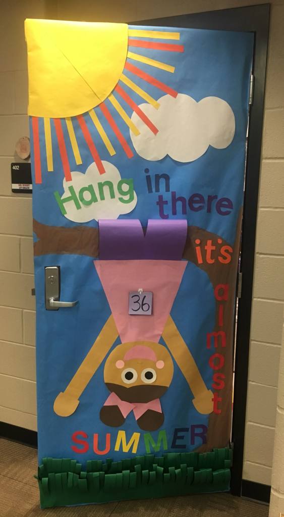 A classroom door decorated with colorful paper featuring a sun, clouds, and a character hanging upside down. The text reads 'Hang in there, it's almost SUMMER' with the number 36 displayed.