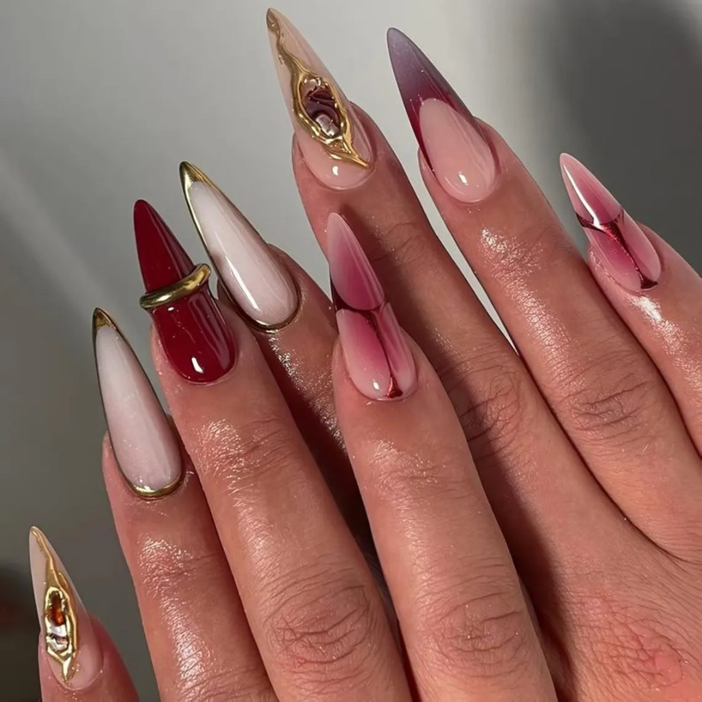 Close-up of beautifully designed long pointed nails featuring a mix of red, pink, and white colors, along with gold accents and intricate patterns.
