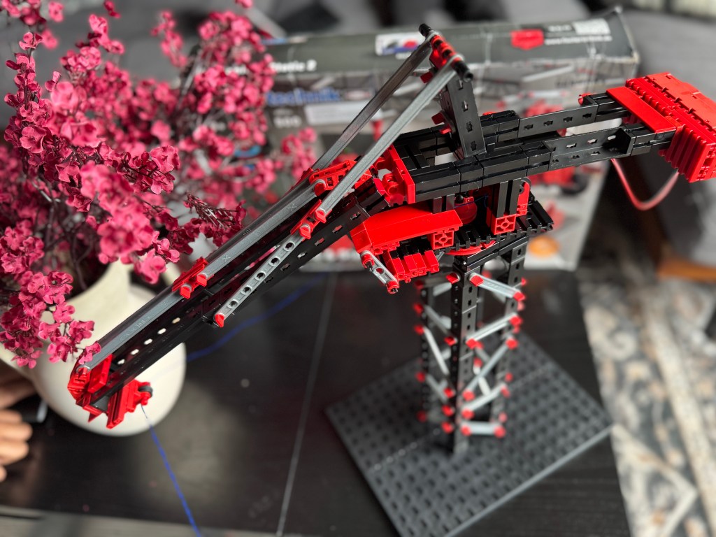 A close-up of a Fischer Technik STEM kit model, featuring a crane structure made of black and red building pieces, alongside a vase of pink flowers.