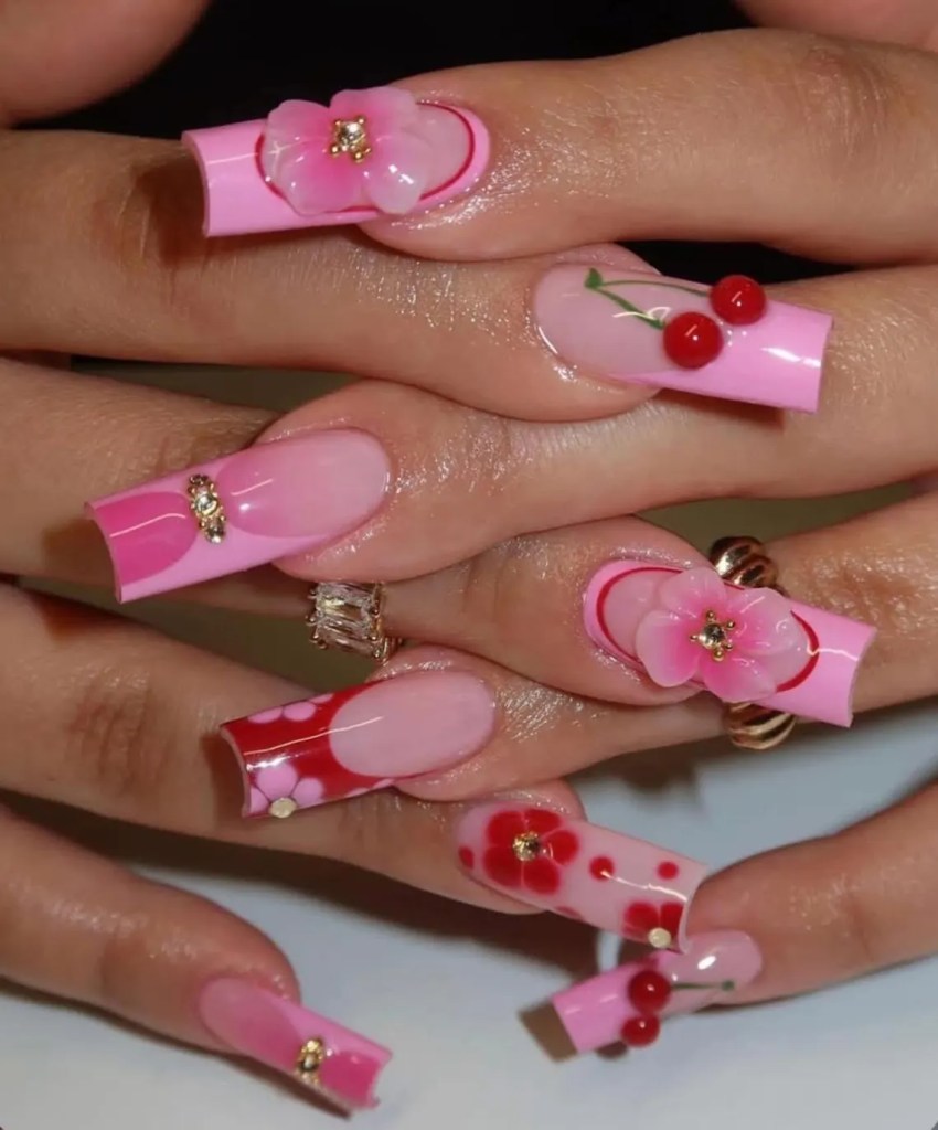 A close-up of beautifully manicured long nails featuring pink polish, floral embellishments, and decorative designs with red accents and gems.