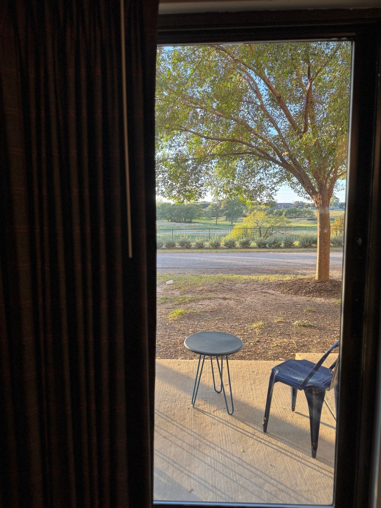 View from inside a room looking out a glass door, featuring a table and a chair on a patio, with a tree and green landscape in the background.
