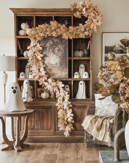 A cozy living space featuring a wooden shelf decorated with ghost figurines, pumpkins, and autumn foliage. A gentle light casts warmth over a comfortable chair adorned with a knitted blanket and soft cushion.