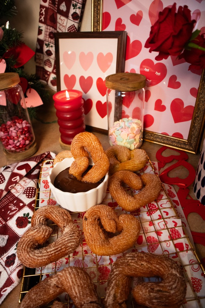 A festive display featuring heart-shaped churros arranged around a bowl of chocolate sauce, with decorative items including a red candle, jars of candies, and heart-patterned artwork.