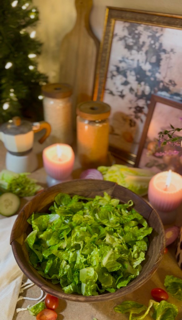 A wooden bowl filled with fresh, chopped romaine lettuce sits on a rustic table, surrounded by candles, decorative jars, and an assortment of vegetables, creating a cozy and inviting atmosphere.