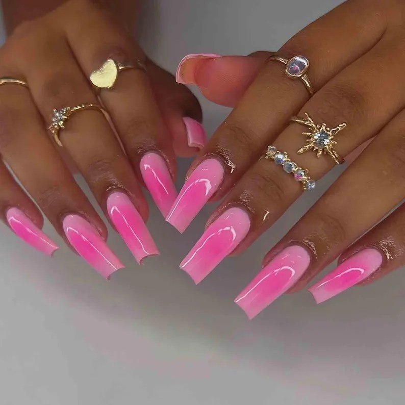 A close-up view of elegant nails with a gradient pink manicure, featuring long stiletto shapes and glossy finish. The hands are adorned with several delicate gold rings.
