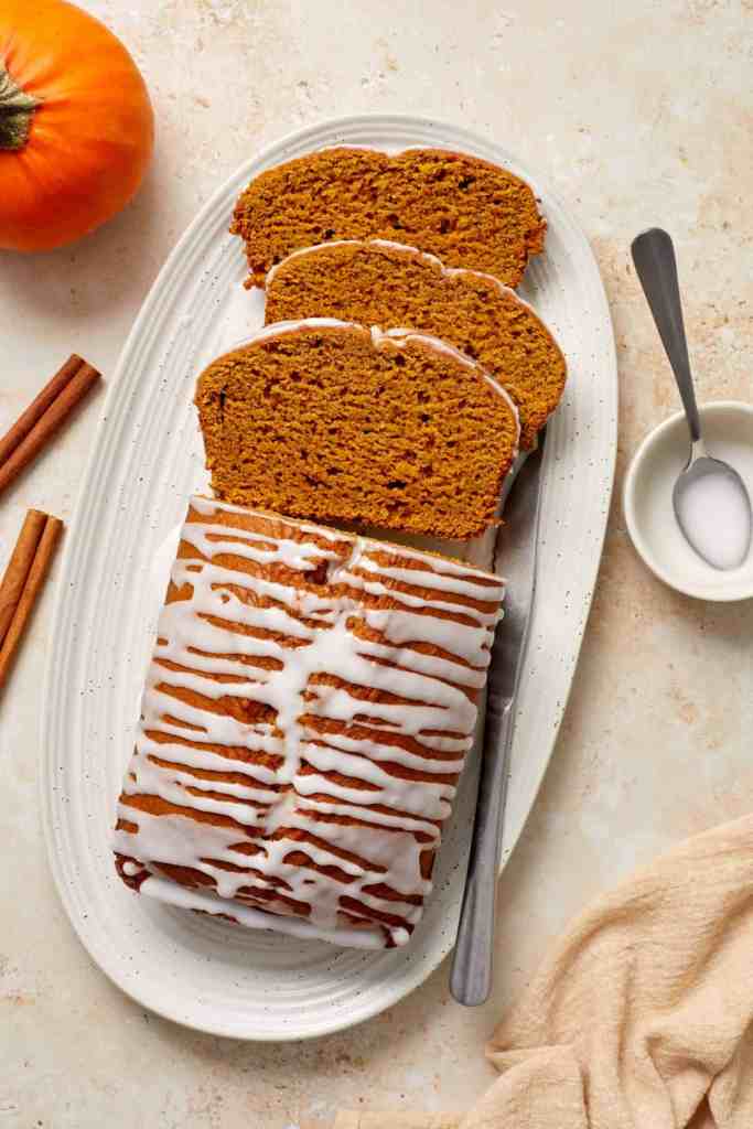 A sliced pumpkin loaf cake drizzled with icing, displayed on a white platter alongside a small bowl of sugar, cinnamon sticks, and a pumpkin.