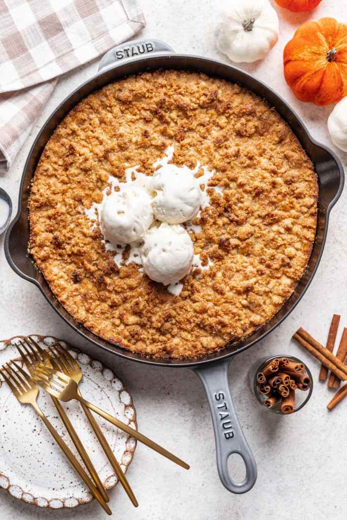 A baked pumpkin crisp dessert topped with three scoops of vanilla ice cream, served in a cast iron dish, with golden forks and cinnamon sticks nearby.