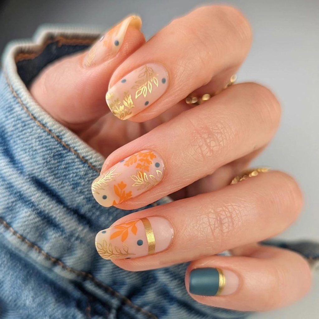 A close-up of manicured nails featuring a transparent base with gold leaf accents, orange floral designs, and blue dots, complemented by a green accent nail with a gold tip.