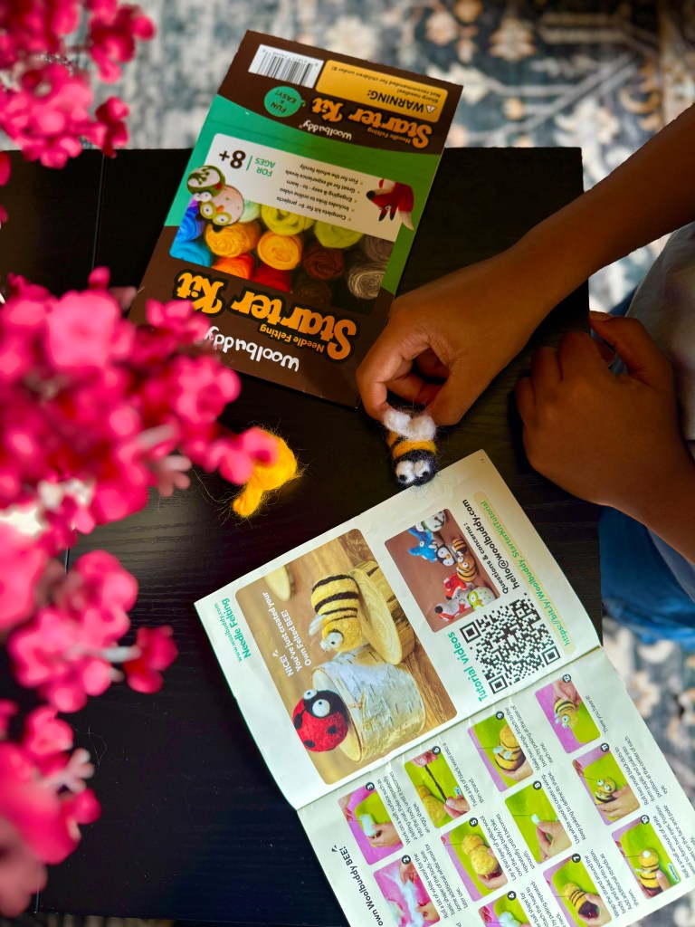 A child holding a needle-felted creature while studying the instruction booklet for the Woolbuddy Needle Felting Starter Kit, with vibrant wool roving colors and blooming flowers in the foreground.