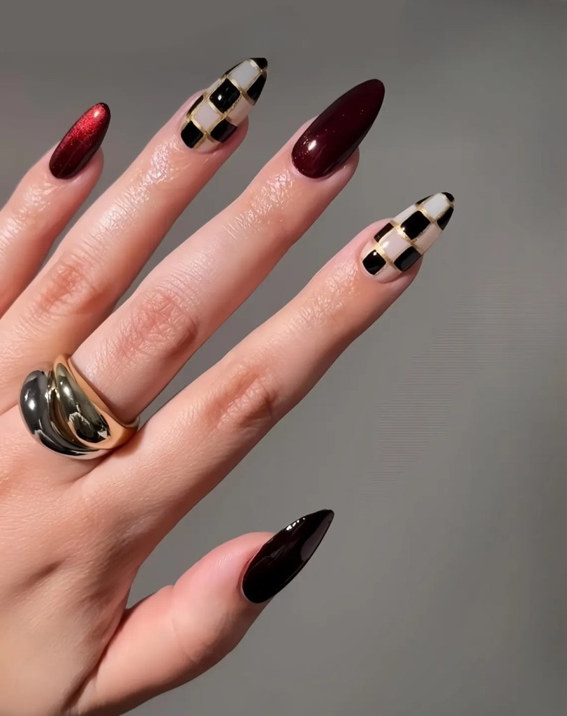 A close-up of a hand with long, pointed nails featuring a mix of dark red and a black-and-white checkered design, complemented by a gold and black ring on the finger.
