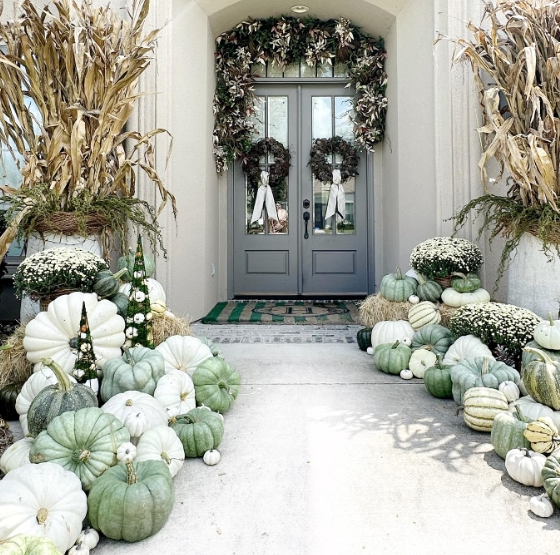 A charming fall porch featuring double gray doors adorned with two wreaths, flanked by autumn foliage. The entrance is decorated with a variety of pale green and white pumpkins arranged across the steps, creating a warm and inviting atmosphere.
