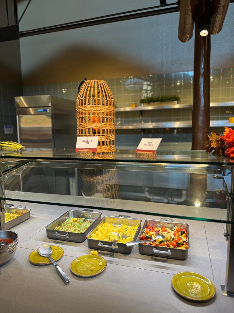 A buffet display featuring scrambled eggs, mixed vegetables, and other breakfast options in a well-lit cafeteria setting.